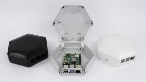 Caja hexagonal para Raspberry Pi, desmontada y con una placa Raspberry Pi instalada en su interior. Se muestran tres versiones del diseño: una carcasa negra cerrada, una carcasa blanca cerrada y una abierta con la placa visible. La caja cuenta con múltiples puertos de conexión y un diseño moderno y funcional.